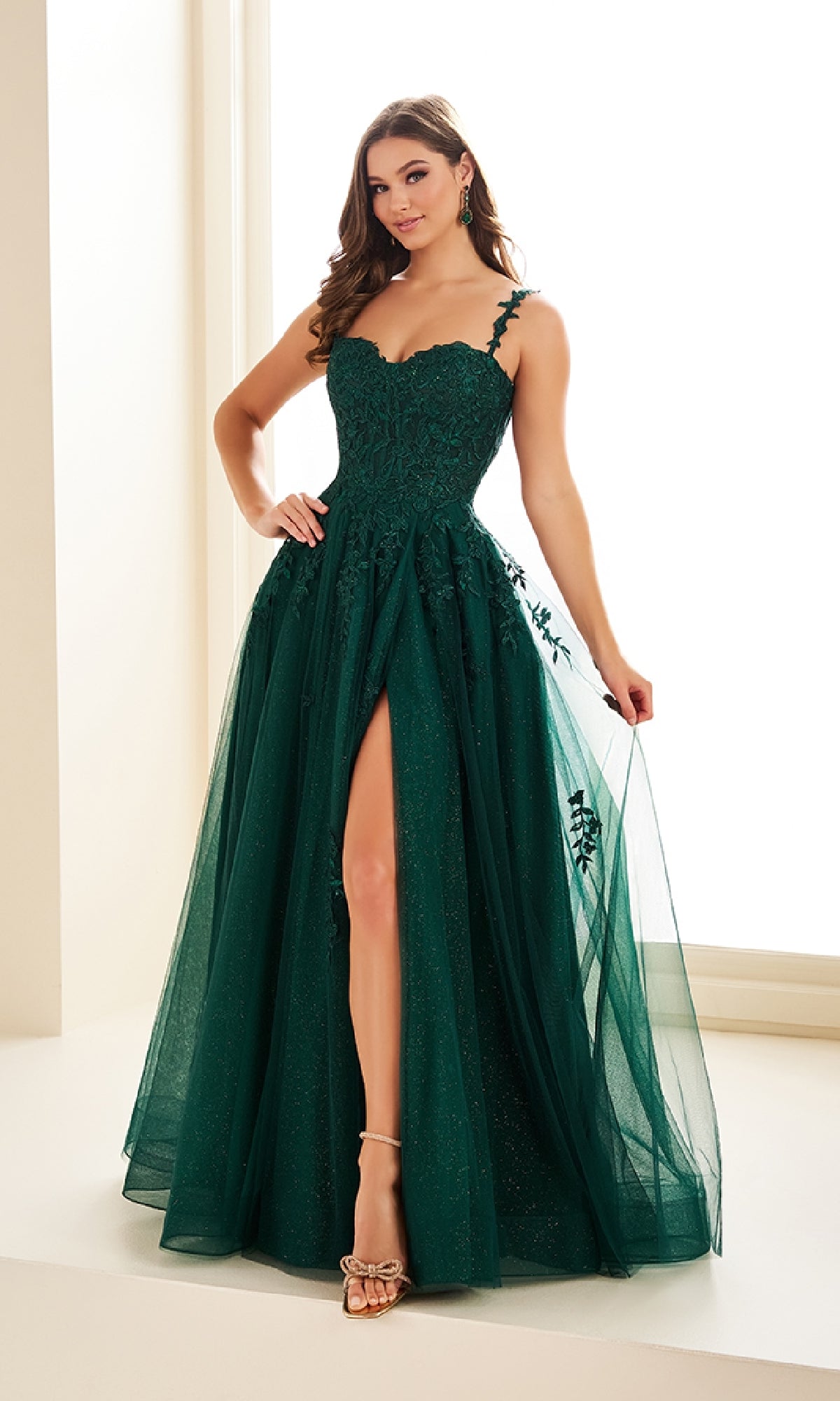 A woman in the Ellie Wilde EW36083 long prom dress, featuring dark green fabric, a sweetheart neckline, and high slit, stands indoors against a light background.